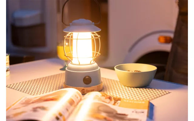 LED фенер осветява маса в къмпинг, заобиколена от чиния и списание, създавайки уютна вечерна атмосфера.