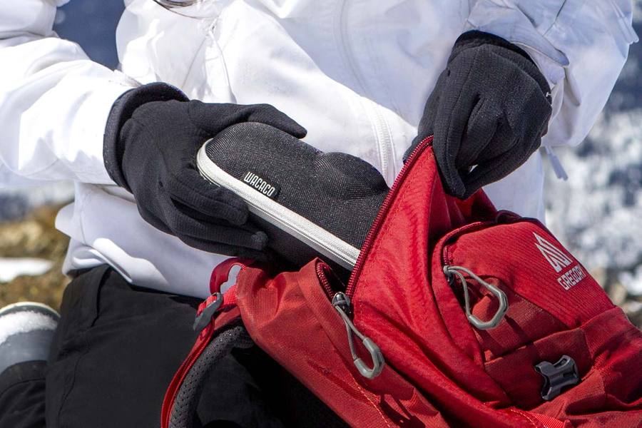 Човек с черни ръкавици поставя предмет в червен раница на фон на снежни планини.