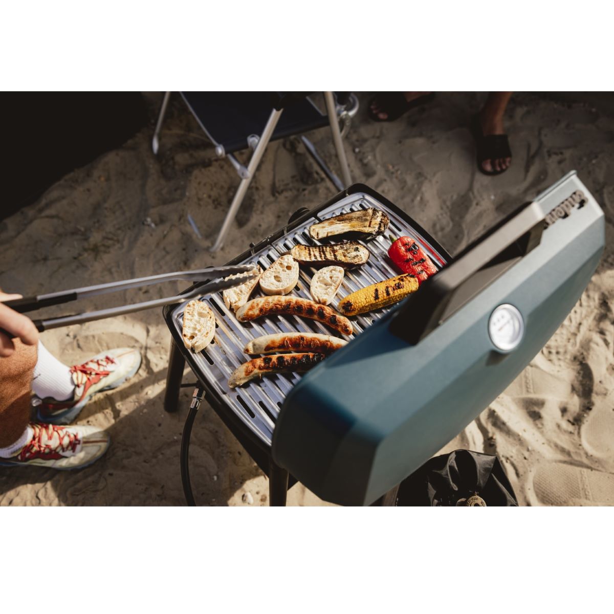 Cooking sausages on Enders Explorer II Pro Caravan gas grill with high stand and shelf on sandy campsite.